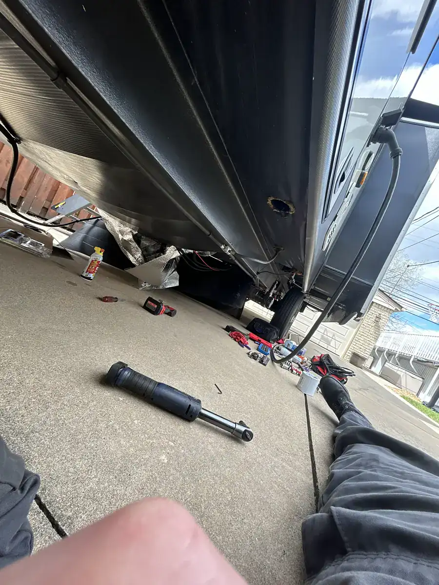 Technician working under an RV on a driveway with power tools and supplies spread on the concrete.