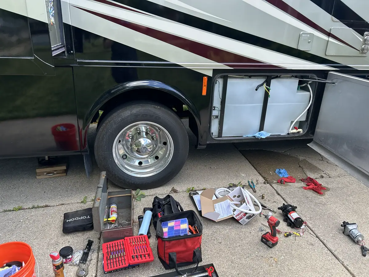 Tools and a new water pump beside an open RV fresh-water tank compartment.